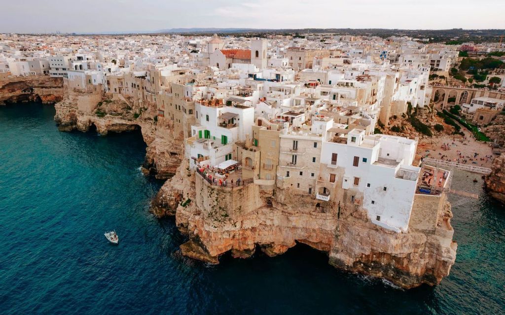 Una veduta aerea di una città con edifici bianchi costruiti su una scogliera rocciosa a picco su un mare turchese.