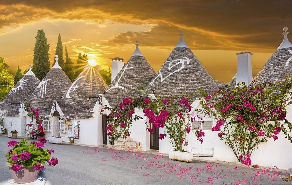 Una fila di trulli bianchi con tetti conici in pietra e fiori rosa lungo una strada sotto un tramonto dorato.
