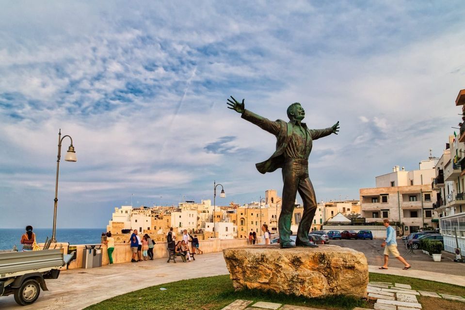 Una statua di bronzo raffigurante un uomo con le braccia tese si erge su una roccia su un lungomare, con una cittadina costiera sullo sfondo.