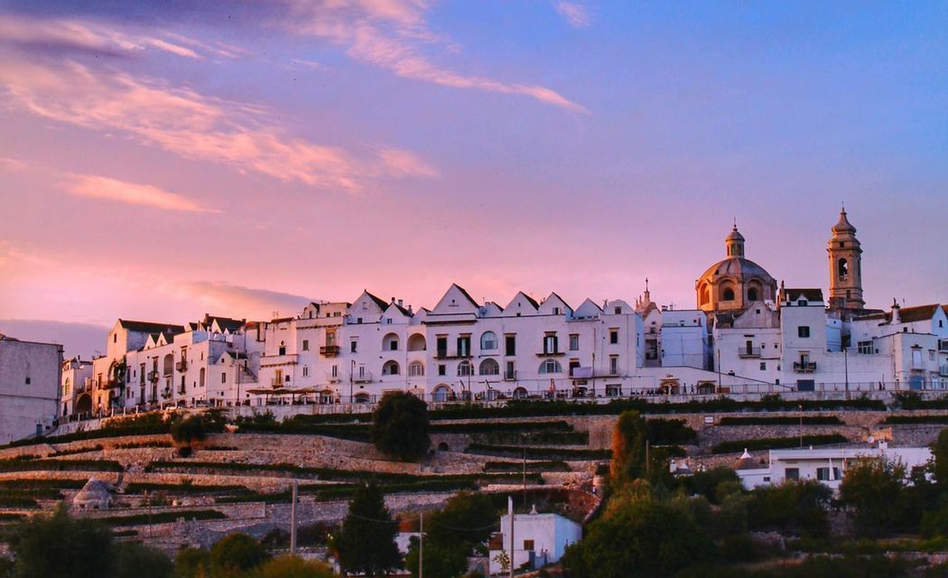 Un paese imbiancato a calce con una chiesa a cupola e un campanile si trova su una collina terrazzata sotto un cielo rosa e blu al tramonto.