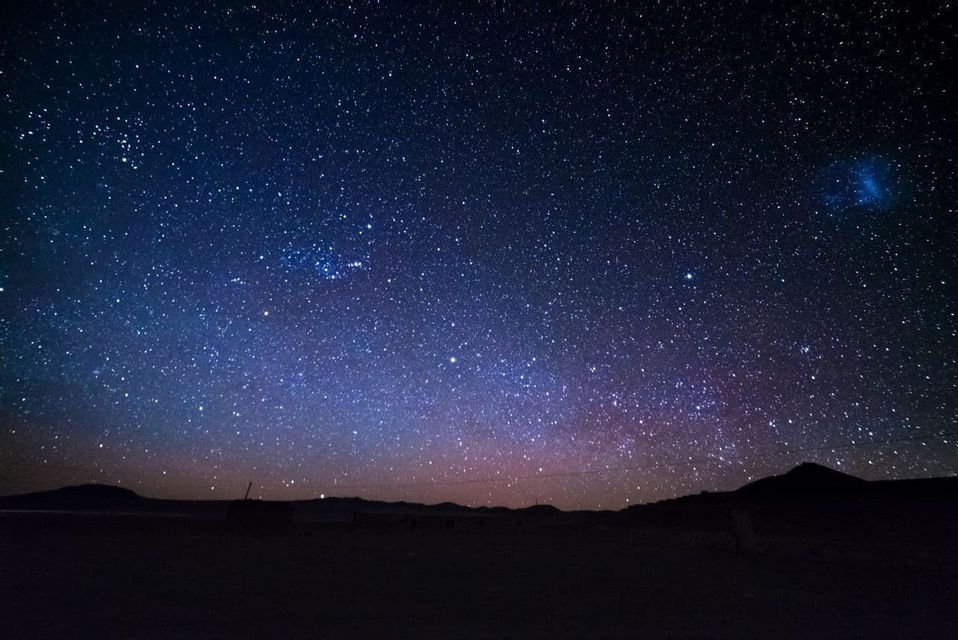 La Via Lattea risplende in sfumature di blu e viola in un vasto cielo notturno stellato sopra un paesaggio in silhouette.