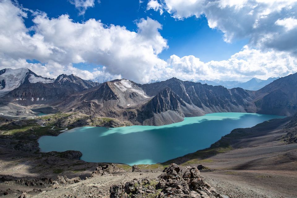 An aerial view of a turquoise alpine lake nestled in a rocky mountain range with snow-capped peaks under a blue, cloudy sky.
