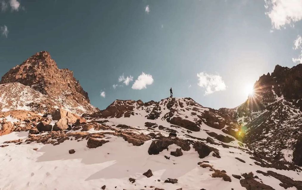 A person stands silhouetted on a snowy mountain peak, looking out at a mountain range under a sunny sky.