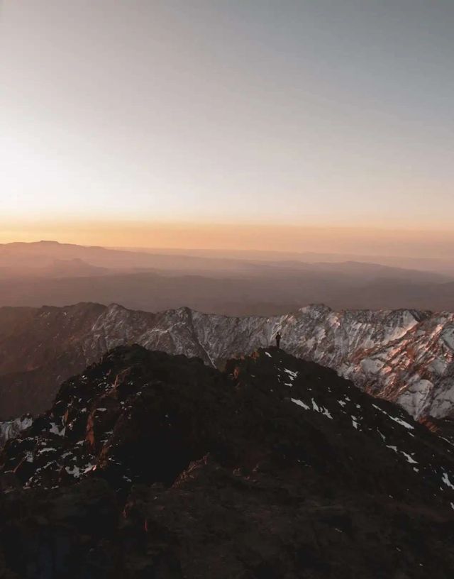A silhouette of a person stands on a mountain peak, viewing a vast mountain range with snow at sunset.