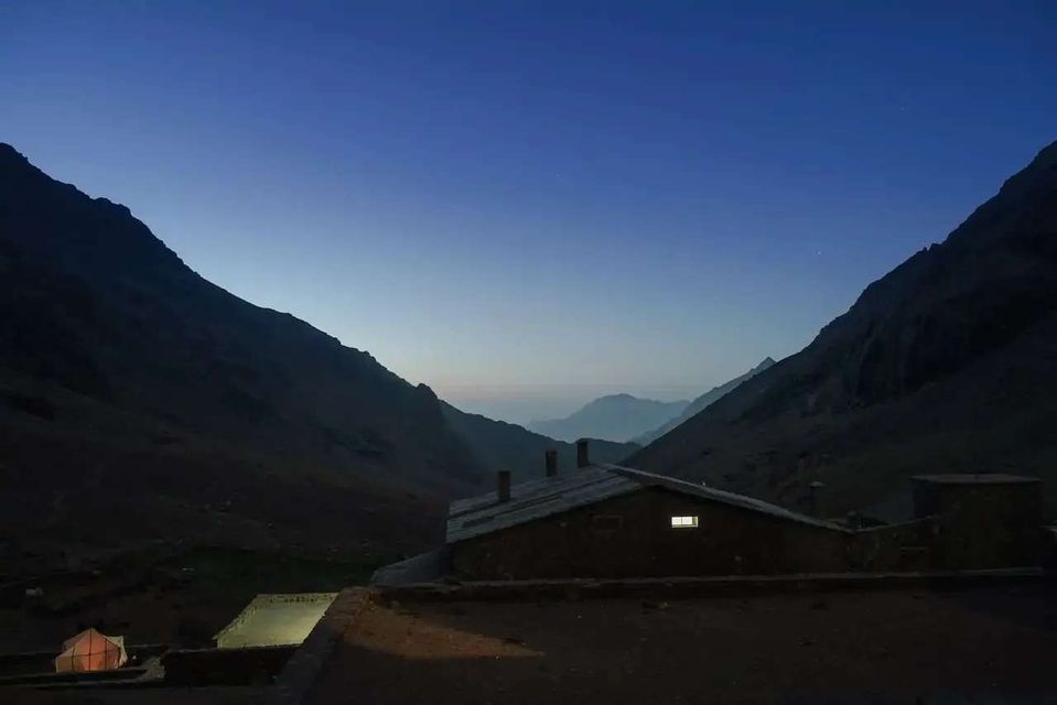 A mountain refuge with a single illuminated window sits in a valley between two dark mountain slopes at dusk.