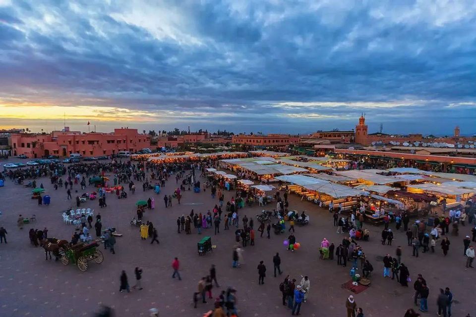 An aerial view of a bustling city square filled with people and illuminated market stalls at dusk under a cloudy sky.