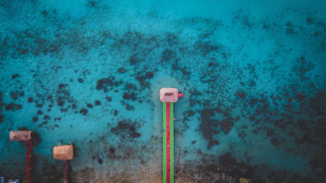 Una vista aerea di un lungo pontile colorato con una capanna alla sua estremità, che si estende su acque limpide color turchese con barriere coralline sottostanti.