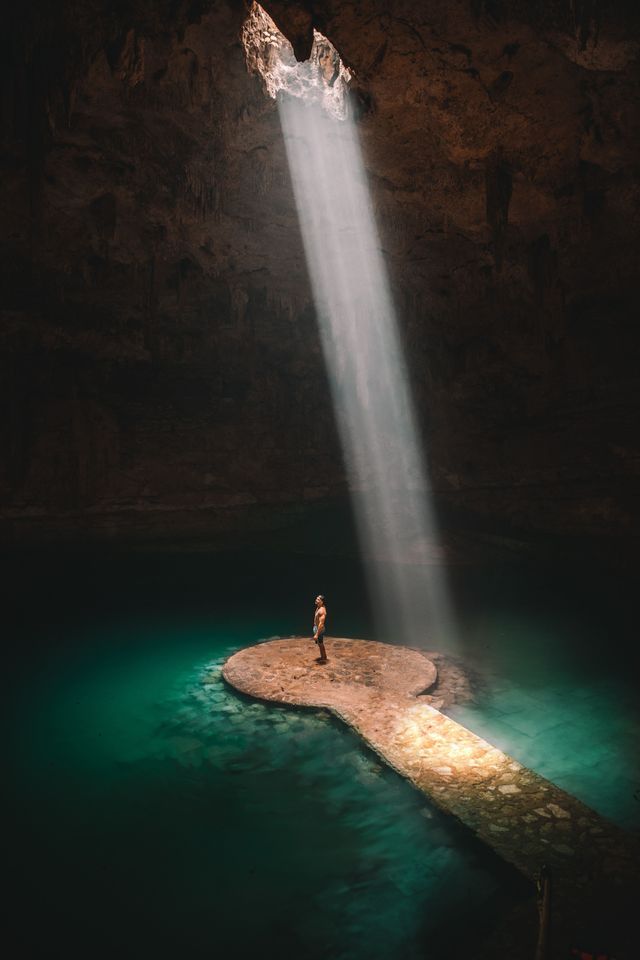 Una persona si trova su una piattaforma di pietra in un cenote, illuminata da un raggio di luce proveniente da un'apertura nel soffitto della grotta.