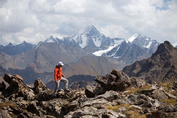 Eine Person in einer orangefarbenen Jacke wandert auf einem felsigen Bergpfad mit schneebedeckten Gipfeln in der Ferne unter einem bewölkten Himmel.