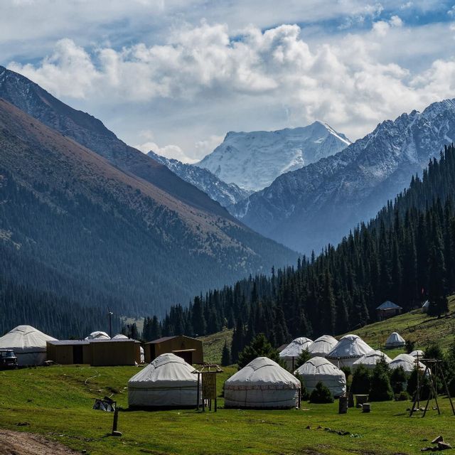 A cluster of traditional white yurts on a green hillside in a valley, with tall, snow-capped mountains in the background.