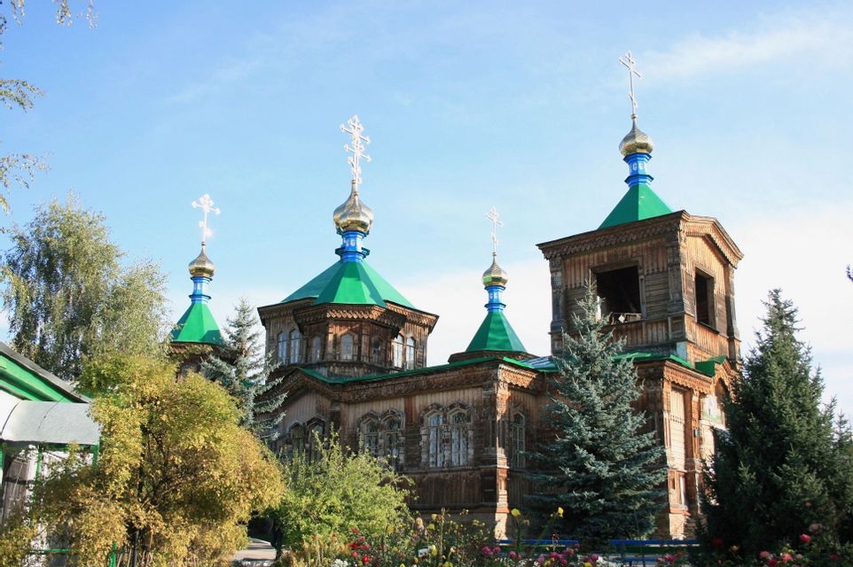 A large wooden Orthodox church with green roofs and golden onion domes surrounded by trees and flowers under a blue sky.