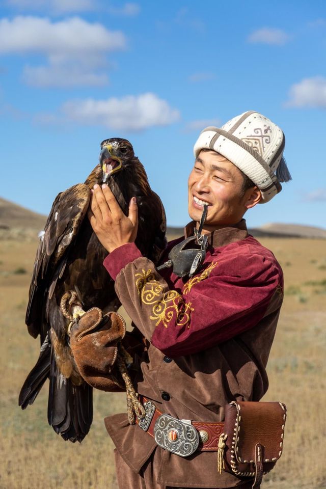Un uomo sorridente in abiti tradizionali tiene una grande aquila reale sulla mano guantata in un campo.