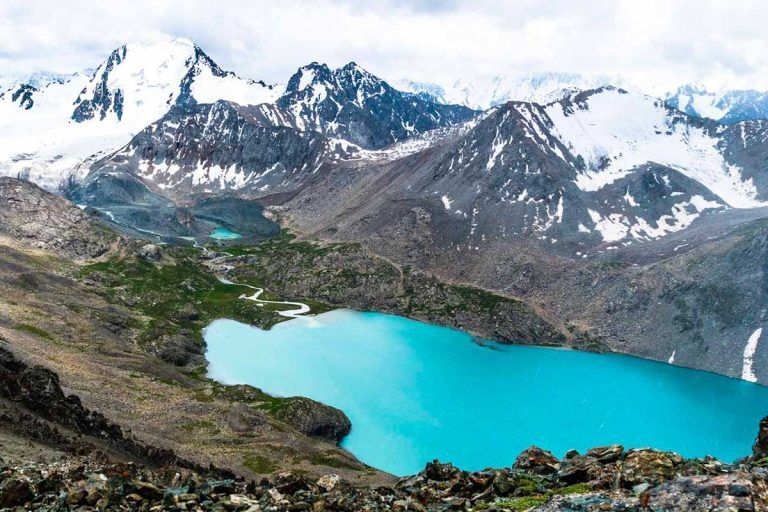 Un lago turchese a forma di cuore giace in una valle rocciosa ai piedi di montagne innevate sotto un cielo nuvoloso.