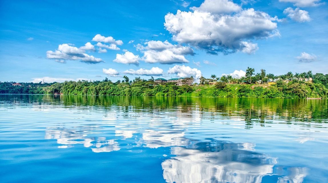 Un lago en calma refleja un cielo azul y nubes blancas hinchadas, con una orilla verde exuberante y edificios visibles en la distancia.