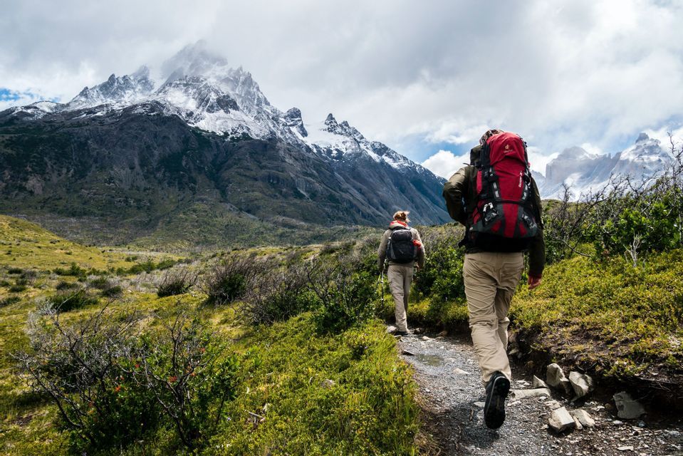 Two hikers with large backpacks walk on a narrow trail through a green valley towards jagged, snow-capped mountains.