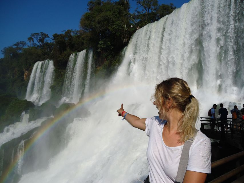 Una mujer rubia con cola de caballo señala un arcoíris que se forma en la neblina de una cascada grande y potente.