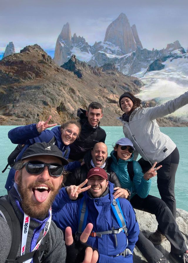 Un grupo de WeRoad se toma una selfie sonriente junto a un lago turquesa con montañas nevadas de fondo.