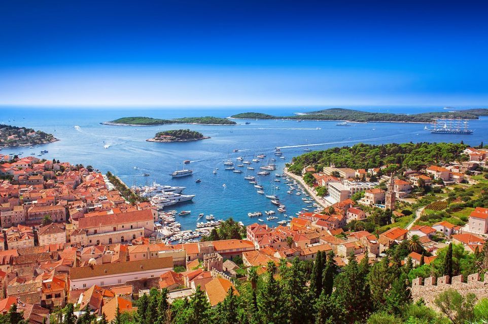 Blick aus der Vogelperspektive auf eine Küstenstadt mit Terrakottadächern, die einen Hafen voller Yachten und Boote überblickt, mit Inseln in der Ferne.