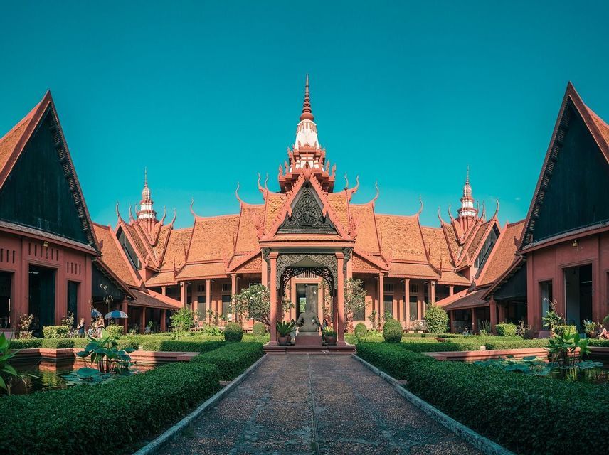 A terracotta-colored building with traditional ornate roofs stands behind a formal garden with a central stone path under a clear blue sky.