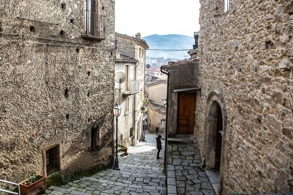 Una persona si trova in uno stretto vicolo acciottolato con gradini, tra vecchi edifici in pietra, con vista sulle montagne in lontananza.