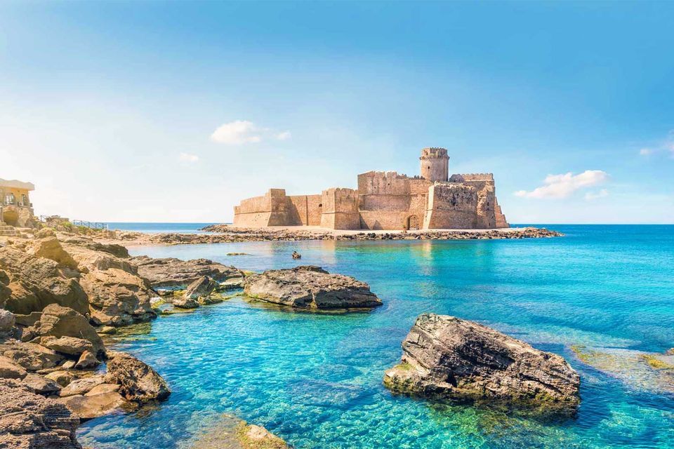 Un castello di pietra sorge su una penisola nel mare cristallino turchese, con una costa rocciosa in primo piano.