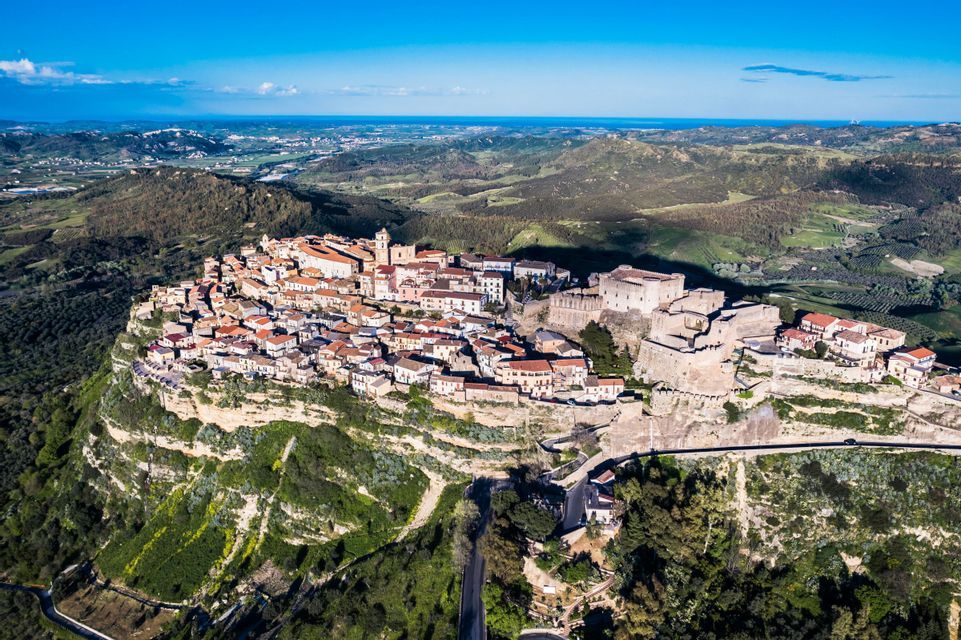 Una vista aerea di una città storica e una fortezza arroccata su una scogliera, che domina un vasto paesaggio verde e il mare in lontananza.