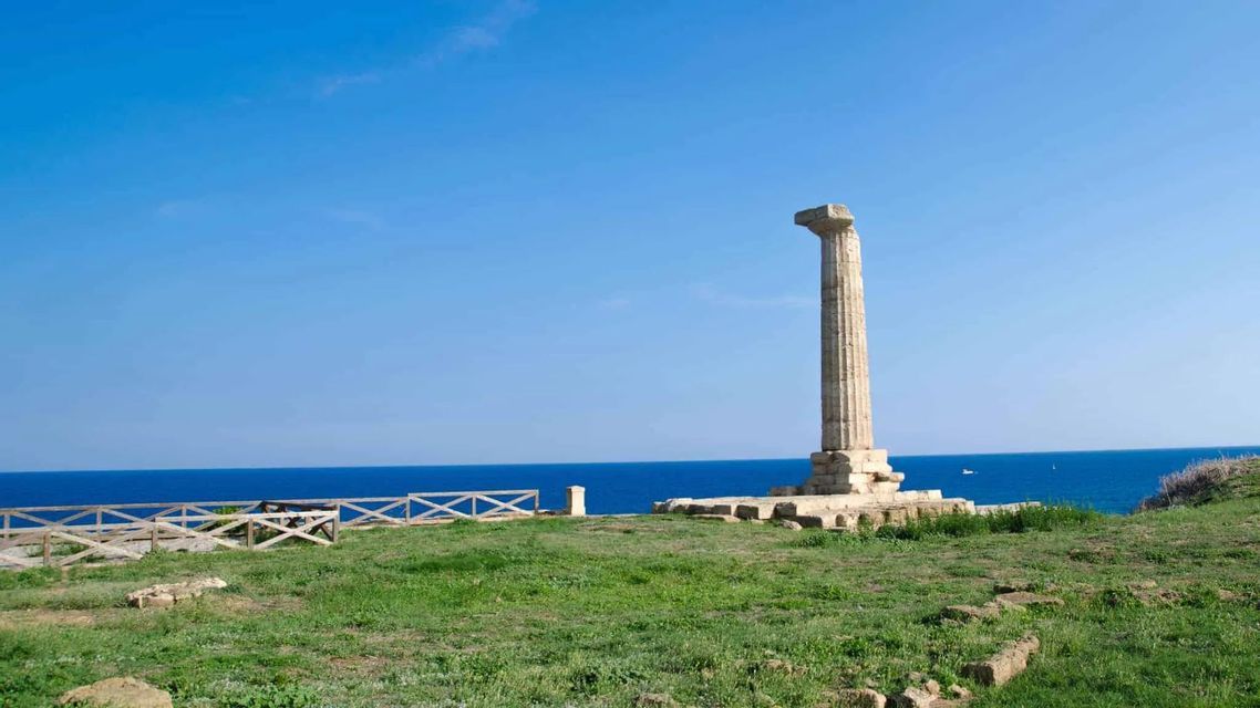 Una colonna antica solitaria su una scogliera erbosa che si affaccia sul mare blu profondo sotto un cielo limpido