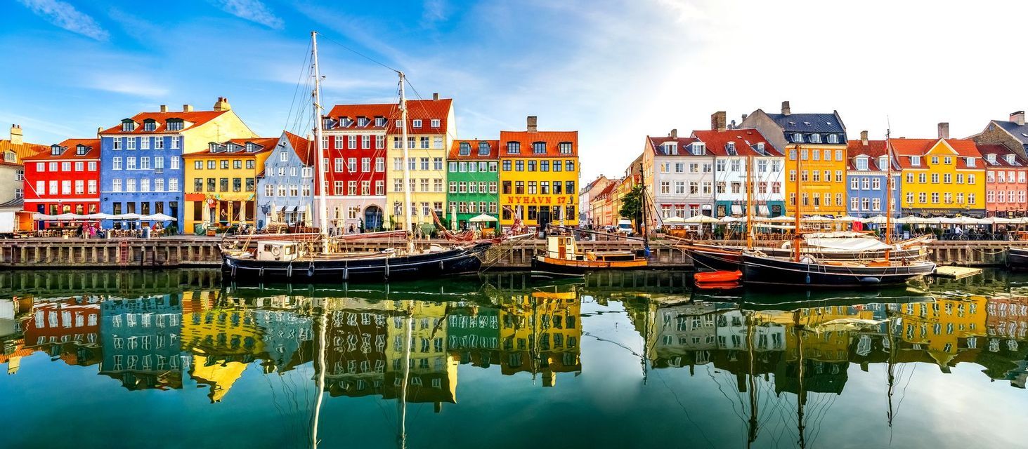 Una hilera de edificios coloridos y barcos amarrados bordean un canal, sus reflejos se espejan en el agua tranquila bajo un cielo azul.
