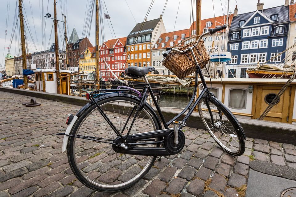 Una bicicleta negra con una cesta de mimbre está aparcada en una calle adoquinada junto a un canal con barcos y edificios coloridos.