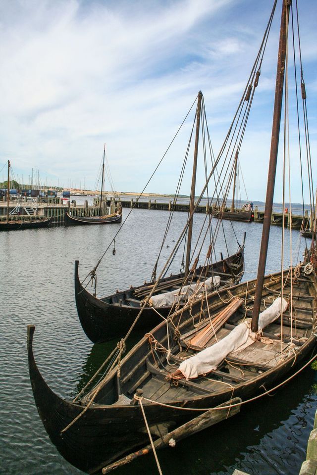 Dos drakkar vikingos de madera oscura, con velas recogidas y mástiles altos, amarrados en un muelle de un puerto.