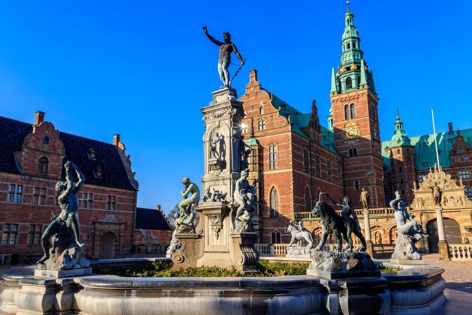 Una fuente ornamentada con estatuas de bronce se encuentra en una plaza frente a un edificio histórico de ladrillo rojo con una torre de reloj.