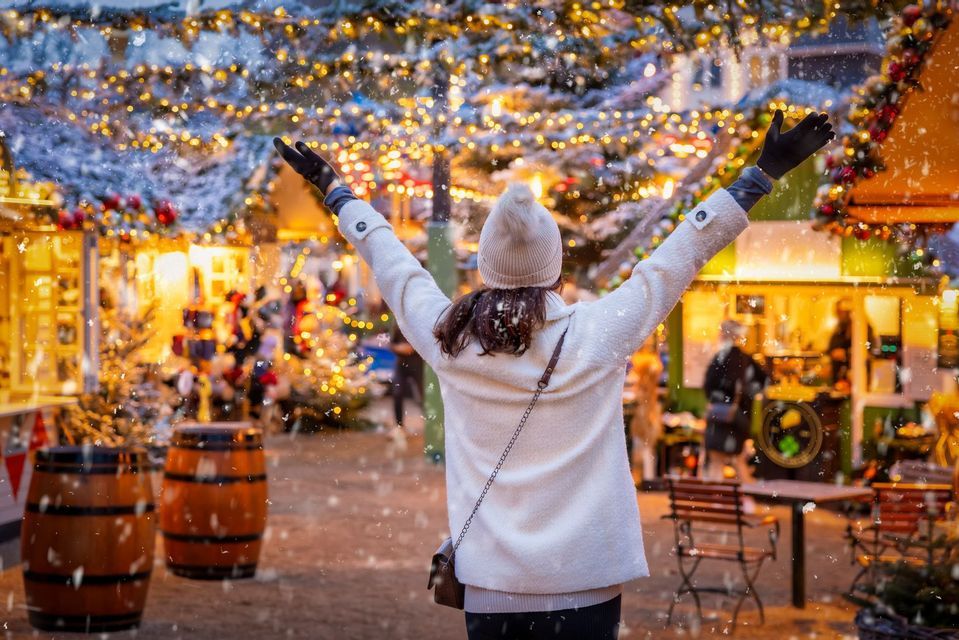 Eine von hinten gesehene Frau hebt ihre Arme auf einem hell erleuchteten Weihnachtsmarkt, während um sie herum Schnee fällt.