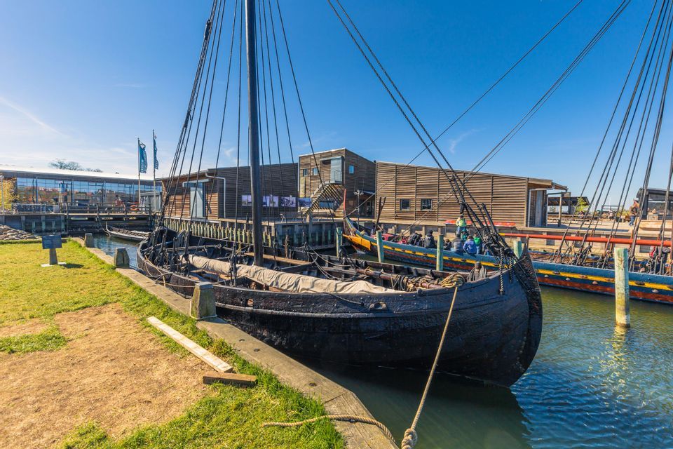 Ein schwarzer Wikingerschiff-Nachbau liegt an einem sonnigen Tag in einem Hafen vor Anker, neben einem Holzsteg und Gebäuden.