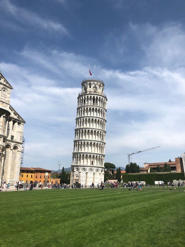 Der Schiefe Turm von Pisa steht auf einer großen grünen Wiese, mit Touristen an seinem Sockel unter blauem Himmel mit weißen Wolken.