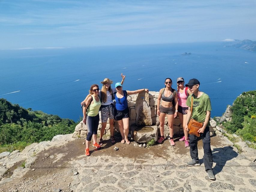 Un groupe WeRoad de six personnes posant pour une photo sur un promontoire rocheux, surplombant une vaste mer bleue par une journée ensoleillée.