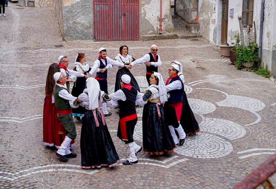 Un gruppo di uomini e donne in abiti tradizionali balla in cerchio, tenendosi per mano su una strada di ciottoli decorata.