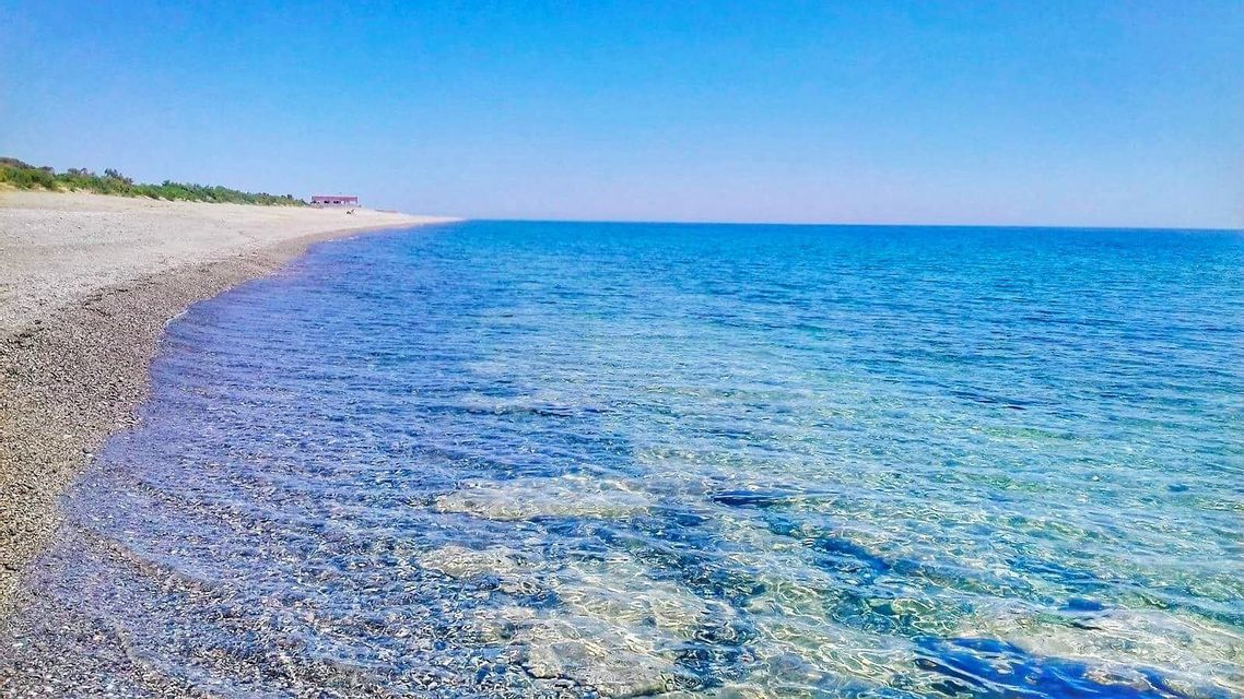 L'acqua limpida e blu lambisce una spiaggia di ciottoli che si estende all'orizzonte sotto un cielo senza nuvole.