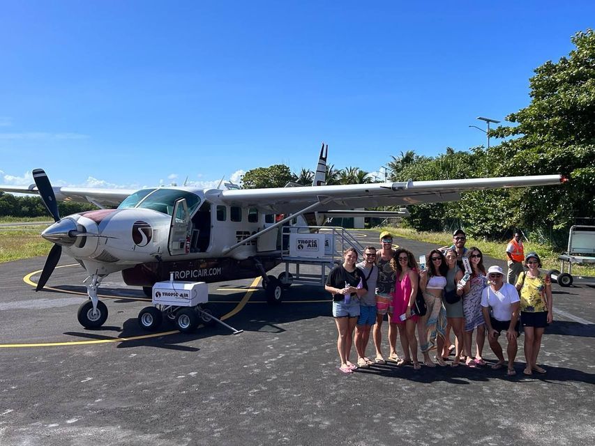 Un gruppo WeRoad posa per una foto sul piazzale di un aeroporto davanti a un piccolo aereo a elica di Tropic Air.