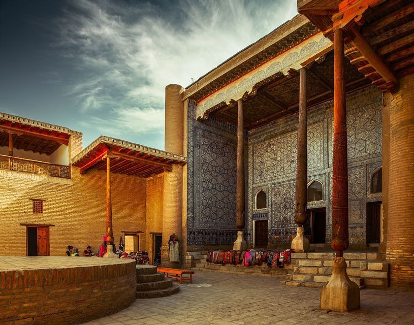 A sunlit courtyard of a historic building with brick walls, a mosaic-tiled facade, and large carved wooden pillars under a cloudy sky.