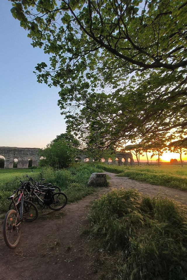 Bicicletas estacionadas en un camino de tierra en un campo de hierba con un acueducto romano al fondo al atardecer.