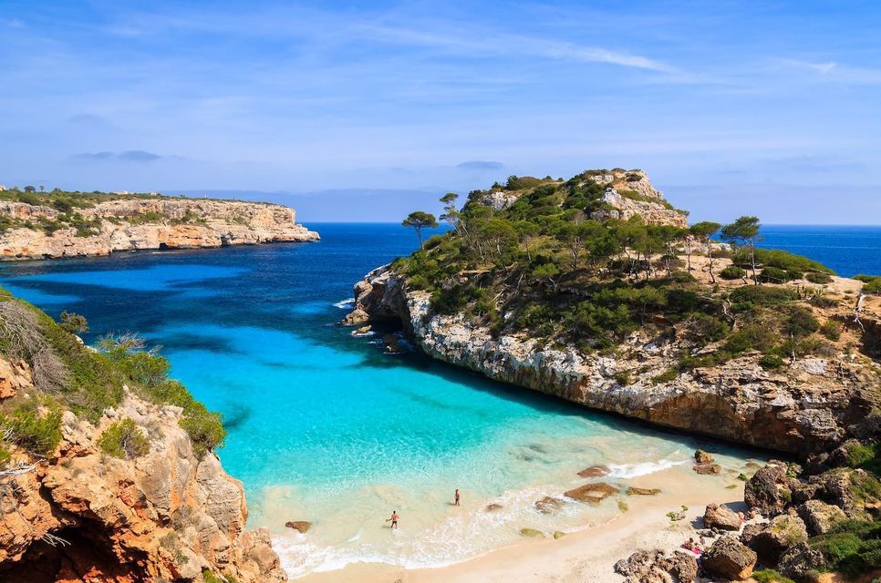 Luftaufnahme einer abgelegenen Bucht mit Sandstrand, klarem türkisfarbenem Wasser und felsigen, baumbewachsenen Klippen.