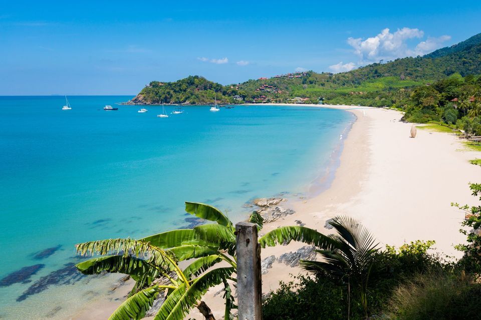 Vue plongeante d'une baie tropicale avec une plage de sable blanc, une eau turquoise claire et des voiliers ancrés au large.