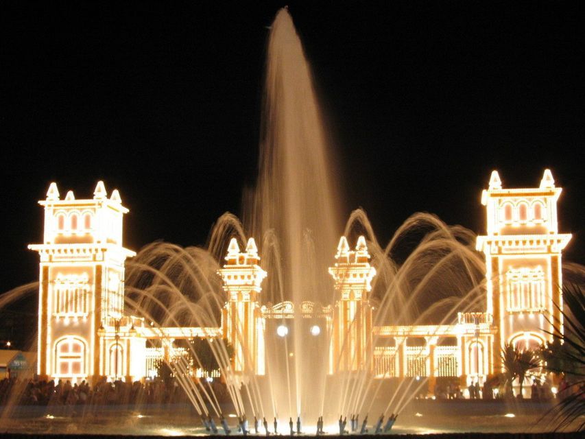 Una grande fontana illuminata spruzza getti d'acqua davanti a un edificio sontuoso decorato con luci di notte.