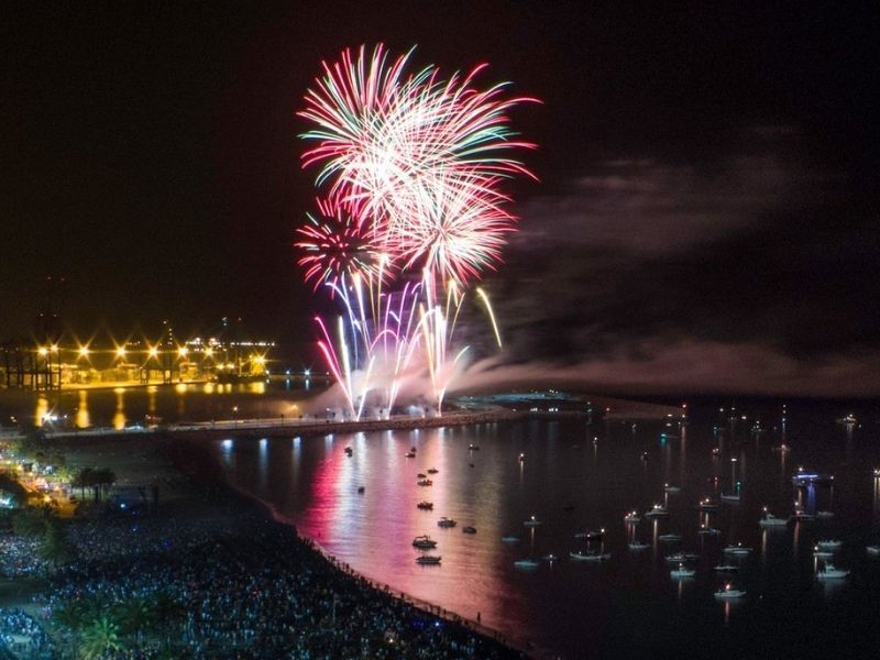 Spettacolari fuochi d'artificio esplodono su un porto di notte, con molte imbarcazioni sull'acqua e una folla che osserva dalla riva.