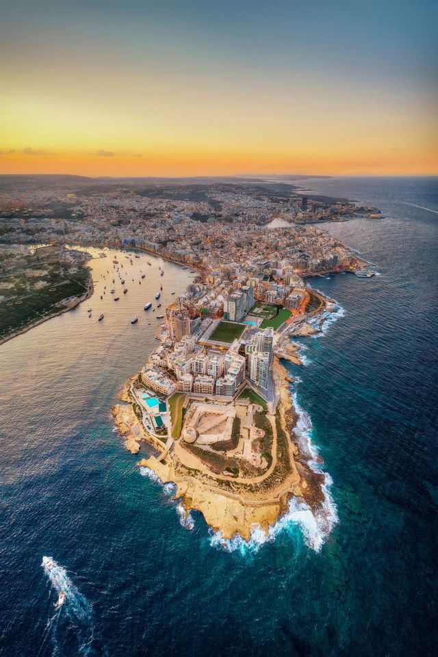 An aerial view of a fortified coastal city on a peninsula, with boats in the harbor under a sunset sky.