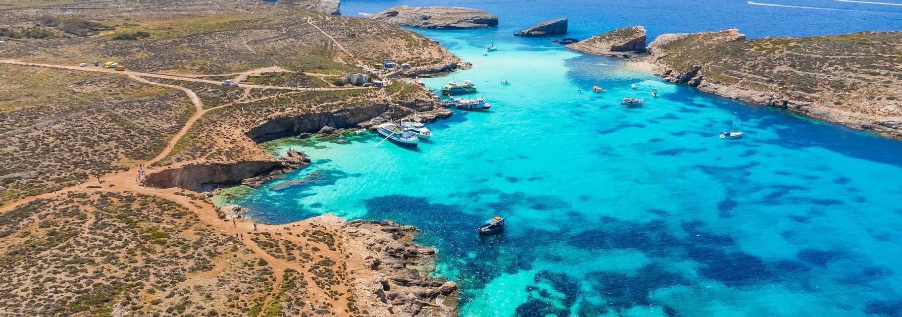 Aerial view of a rocky inlet with vibrant turquoise water, where several boats are docked and people are swimming.