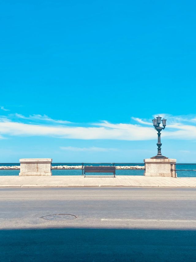 Una panchina vuota e un lampione su un lungomare, con l'oceano visibile sotto un cielo azzurro e limpido.