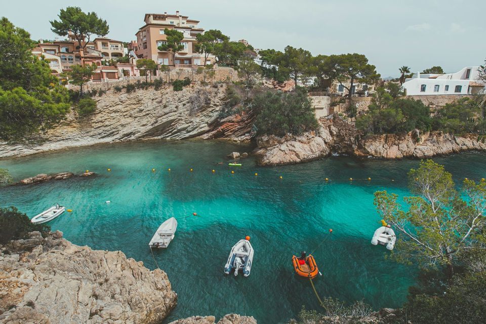 Vista aérea de varias embarcaciones pequeñas amarradas en una cala tranquila y turquesa, rodeada de acantilados rocosos y casas.