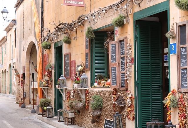 La fachada rústica de un restaurante decorada con persianas verdes, pizarras y pimientos secos colgando en una calle estrecha.