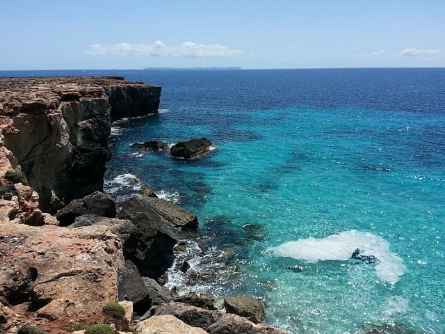 Un nadador en aguas turquesas cristalinas junto a un acantilado rocoso y escarpado bajo un cielo azul.